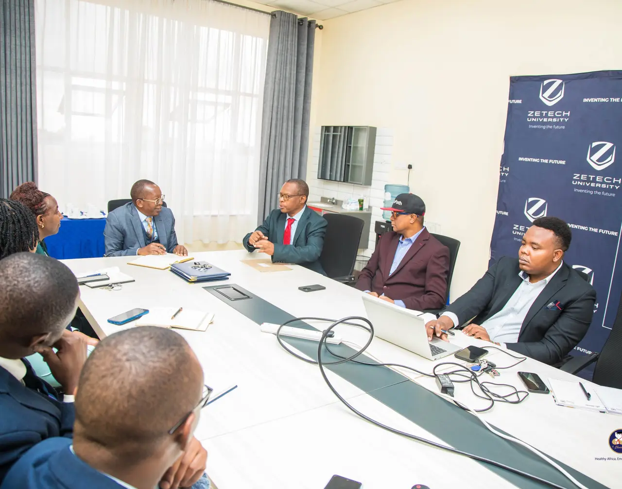 Delegates from I Choose Life – Africa, Zetech University and JuaPath in a working session around the table, with the Zetech University banner in the background.